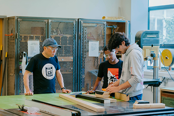 Lecturer Kevin Chen observes two students measuring small blocks of wood in the Wood Shop in the Fine Arts building