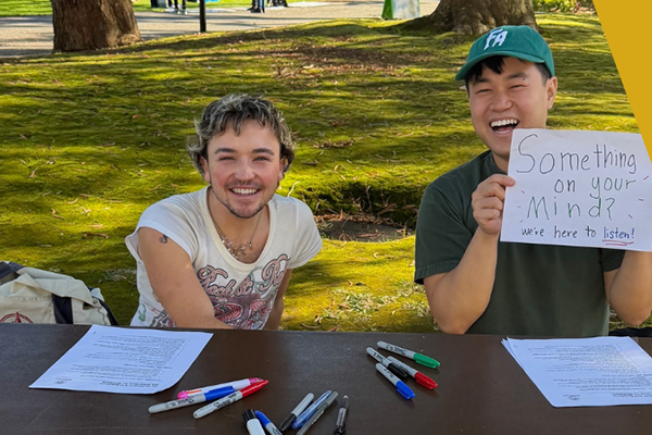 Two students sit at a table outside the student center, smiling and holding a sign that reads, ‘Something on your mind? We’re here to listen.’