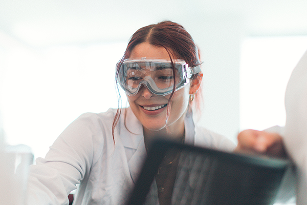 Close-up, worm's-eye view of a smiling student in a lab coat and goggles, observing an experiment or demonstration in a bright white classroom at the Science and Engineering Innovation Center. The foreground is out of focus.