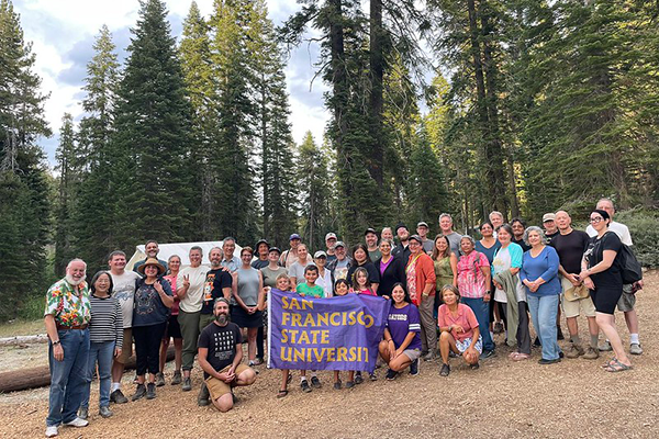 Alumni proudly hold an SFSU flag at a campsite among redwoods