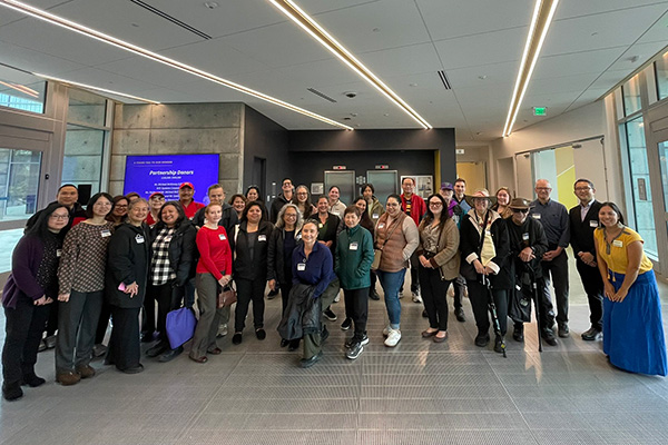 About 20 attendees of the alumni event in the Science & Engineering Innovation Center pose for a photo while standing in a lobby with a large display monitor behind them