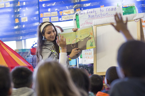 An SFSU student reads aloud from a picture book to a group of children, several of whom are raising their hands.