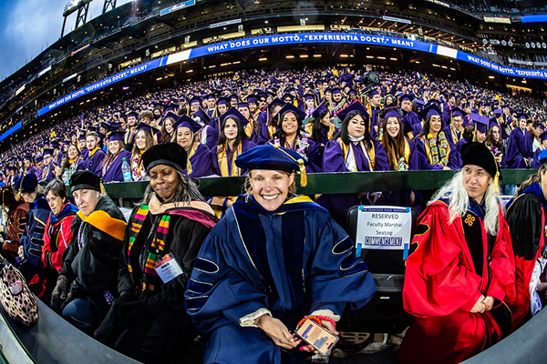 SFSU graduates, wearing purple caps and gowns, sit in the audience at Oracle Park stadium. Faculty members in academic regalia are seated in the front row.