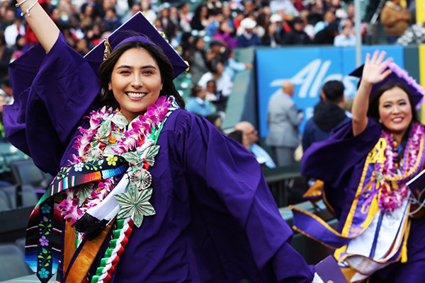 A graduate waves while walking on the warning track at Oracle Park during the Commencement processional