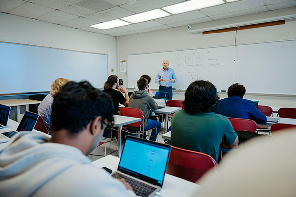 Students use laptops while an instructor presents at the front of a classroom.
