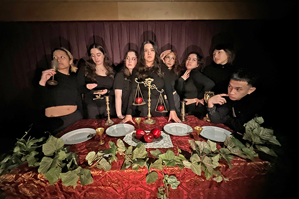 A group of seven dancers dressed in black, standing in front of a table set for a meal.