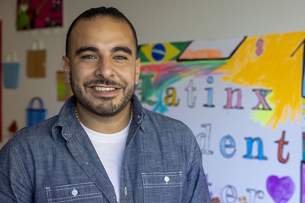 Emmanuel Padilla smiles indoor in front of a hand-drawn Latinx Student Center poster on the wall