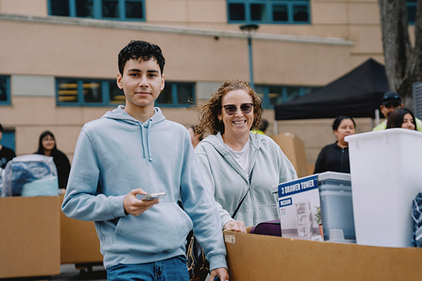 A new SFSU student and their parent push a cart of belongings toward a residence hall on move-in day.