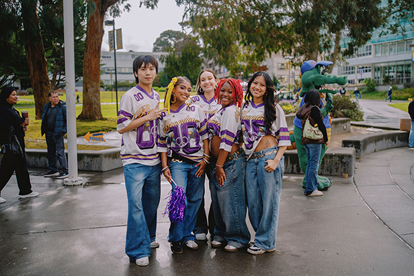 Five people wearing SF State Gators football jerseys and blue jeans stand on Malcolm X Plaza on an overcast and rainy day.