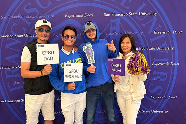 An SFSU student and family pose in front of a step-and-repeat backdrop holding “SFSU Mom,” “SFSU Dad,” “SFSU Brother” and Alli Gator signs.