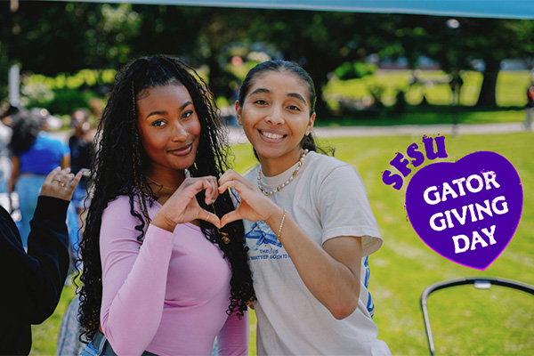 Two SFSU students smile and form a heart shape with their hands with the SFSU Gator Giving Day heart logo displayed to the right.