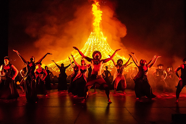 Dancers in traditional Hawaiian dress pose in front of a large fire
