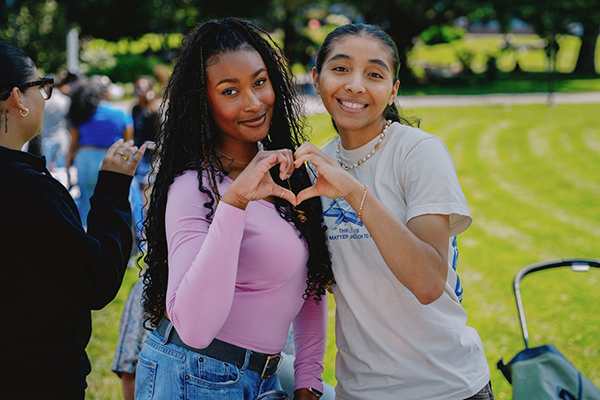 Two SFSU students smile while forming a heart shape with their hands on the sunny campus quad.