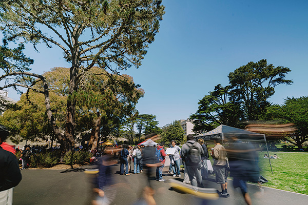 A walking path on campus with a tree line on the left and moving students walking through the middle path