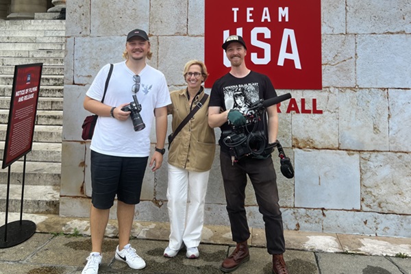 A woman stands with two men in front of a banner saying "Team USA"