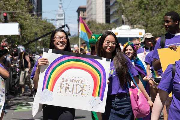 SF State students march in the 2019 San Francisco Pride Parade with two holding a hand-painted sign with a rainbow above the text SF State PRIDE