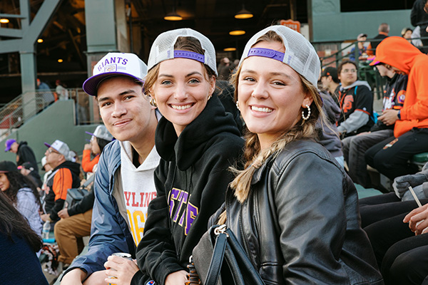 Three people smile while seated and wearing co-branded SFSU-San Francisco Giants baseball caps at Oracle Park.