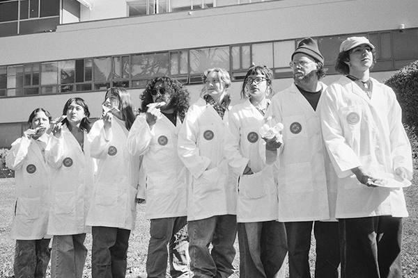 Eight members of the Office of the Ordinary, each wearing a white lab coat with the official seal on the right breast pocket, looking in the distance. The Lam Family College of Business building is visible in the background.