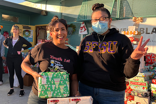 Two people pose for a photo while standing and holding wrapped Christmas presents