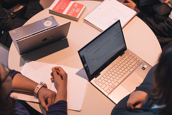 Overhead view of students around a table, using on laptops and writing notes