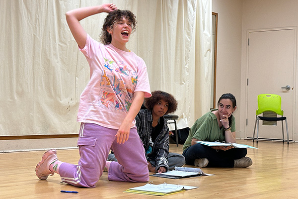 Three actors rehearse a scene on a theater floor; one kneels with an arm raised while two others sit nearby.